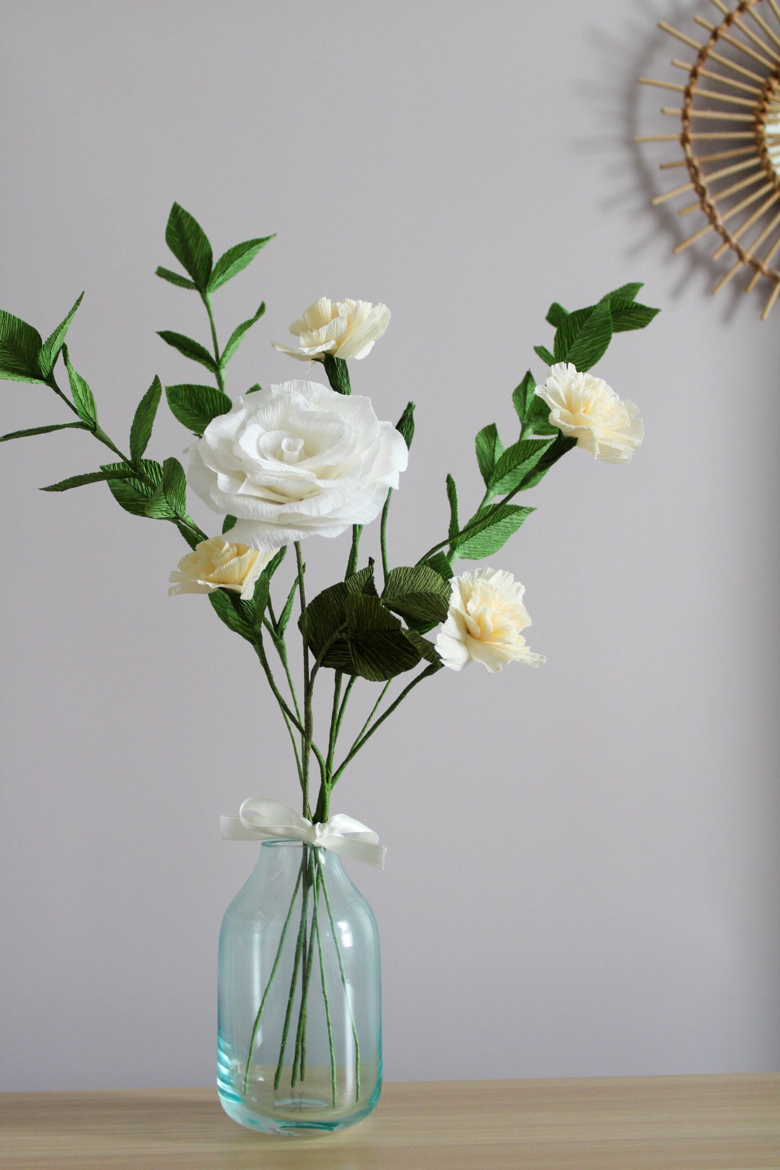 Bouquet de fleurs en papier composé d’une rose, d’œillets spray et de branche de ruscus. Ce bouquet est posé sur un bureau et contenu dans un vase en verre transparent. À l’arrière plan, un mur taupe et une partie d’un miroir en rotin.